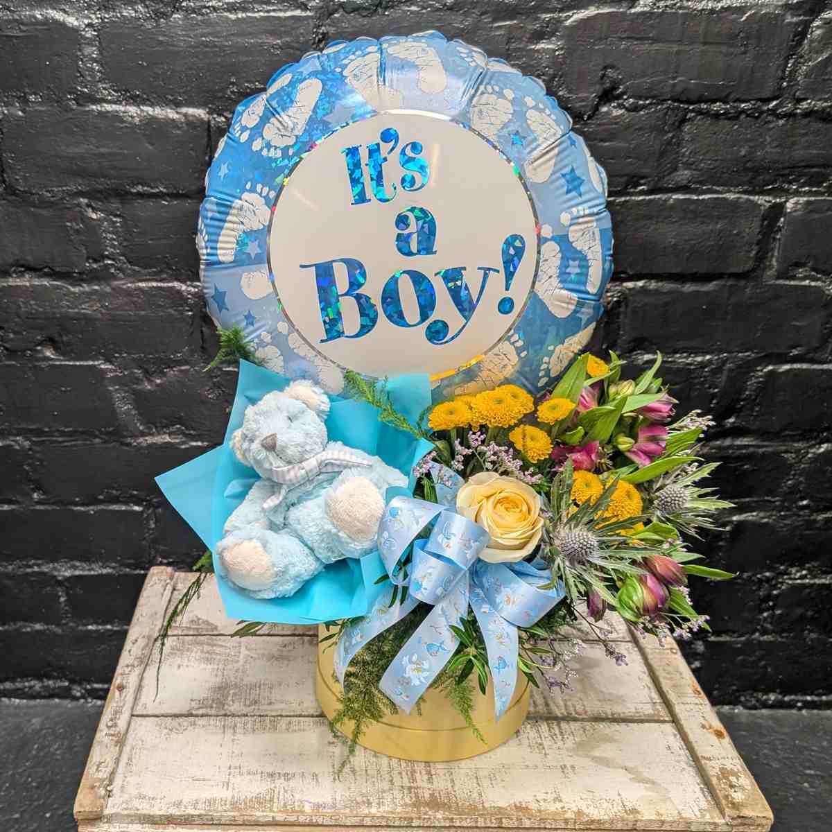 Baby Boy Gift Set “It’s a Boy” new baby gift with blue balloon, soft blue teddy bear and yellow and purple flower arrangement in a gold hat box on a wooden crate – Anastasia Florists Aberdeen - Side View