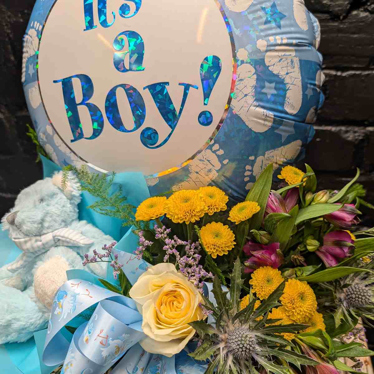 Baby Boy Gift Set “It’s a Boy” new baby gift with blue balloon, soft blue teddy bear and yellow and purple flower arrangement in a gold hat box on a wooden crate – Anastasia Florists Aberdeen - close up