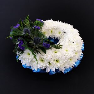 Miniature white chrysanthemum funeral wreath with blue thistles, purple flowers and blue ribbon on a black background