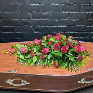 Pink rose and red carnation single ended funeral spray with yellow filler flowers and greenery on a black background.