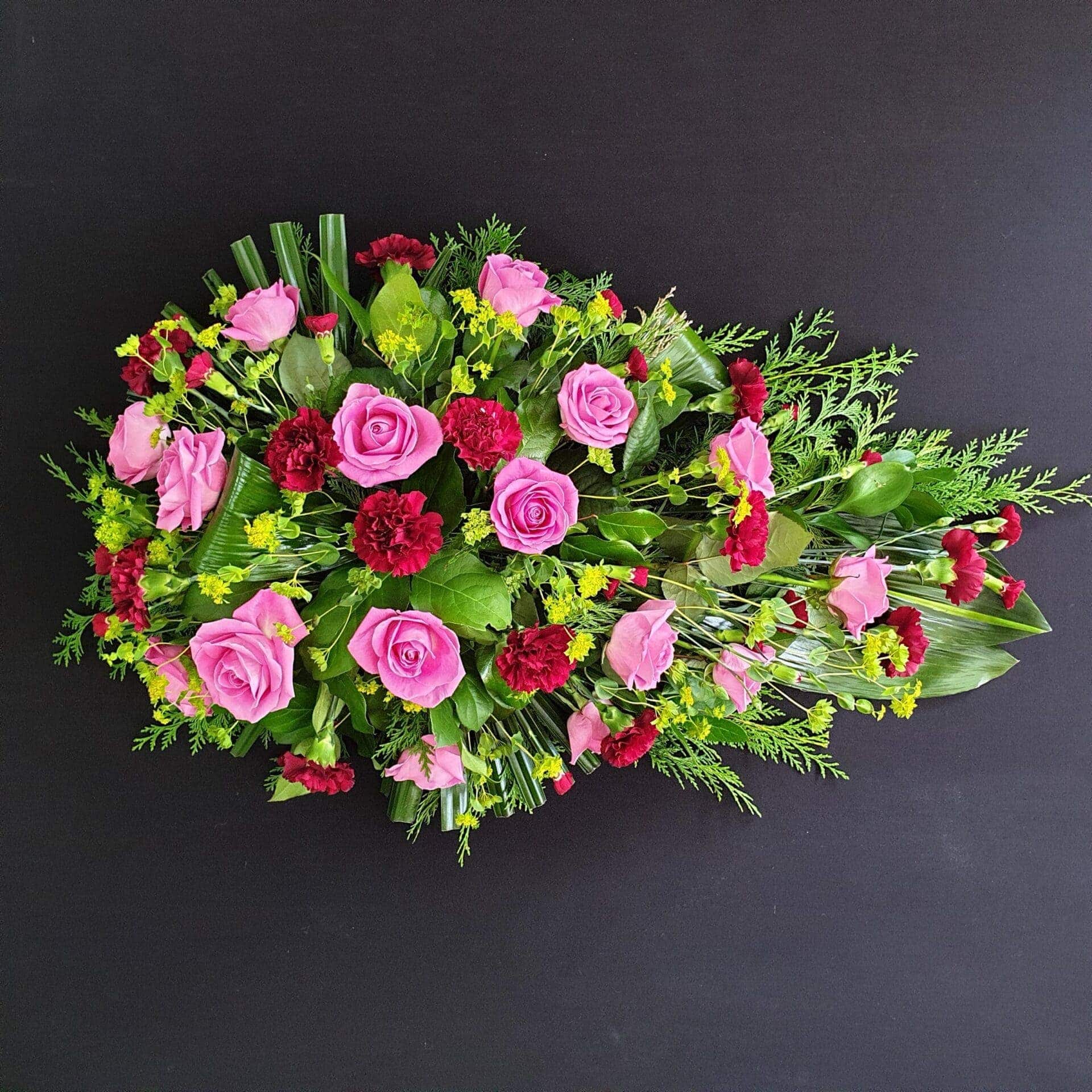 Pink rose and red carnation single ended funeral spray with yellow filler flowers and greenery on a black background.