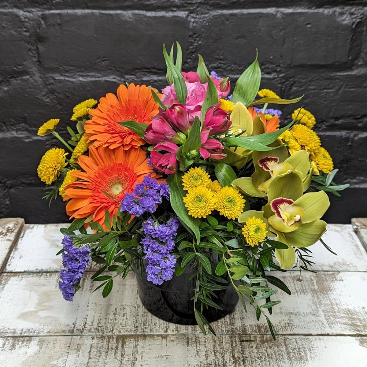 Bright flower arrangement with orange gerberas, yellow chrysanthemums, pink flowers, green orchids and purple blooms in a black container – Anastasia Florists Aberdeen