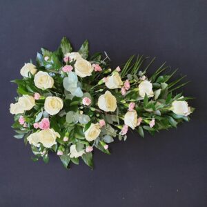 White rose and pale pink carnation single ended funeral spray with eucalyptus and greenery on a dark background.