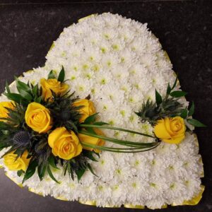White chrysanthemum heart funeral tribute with yellow roses, thistle and yellow ribbon edging on a dark surface