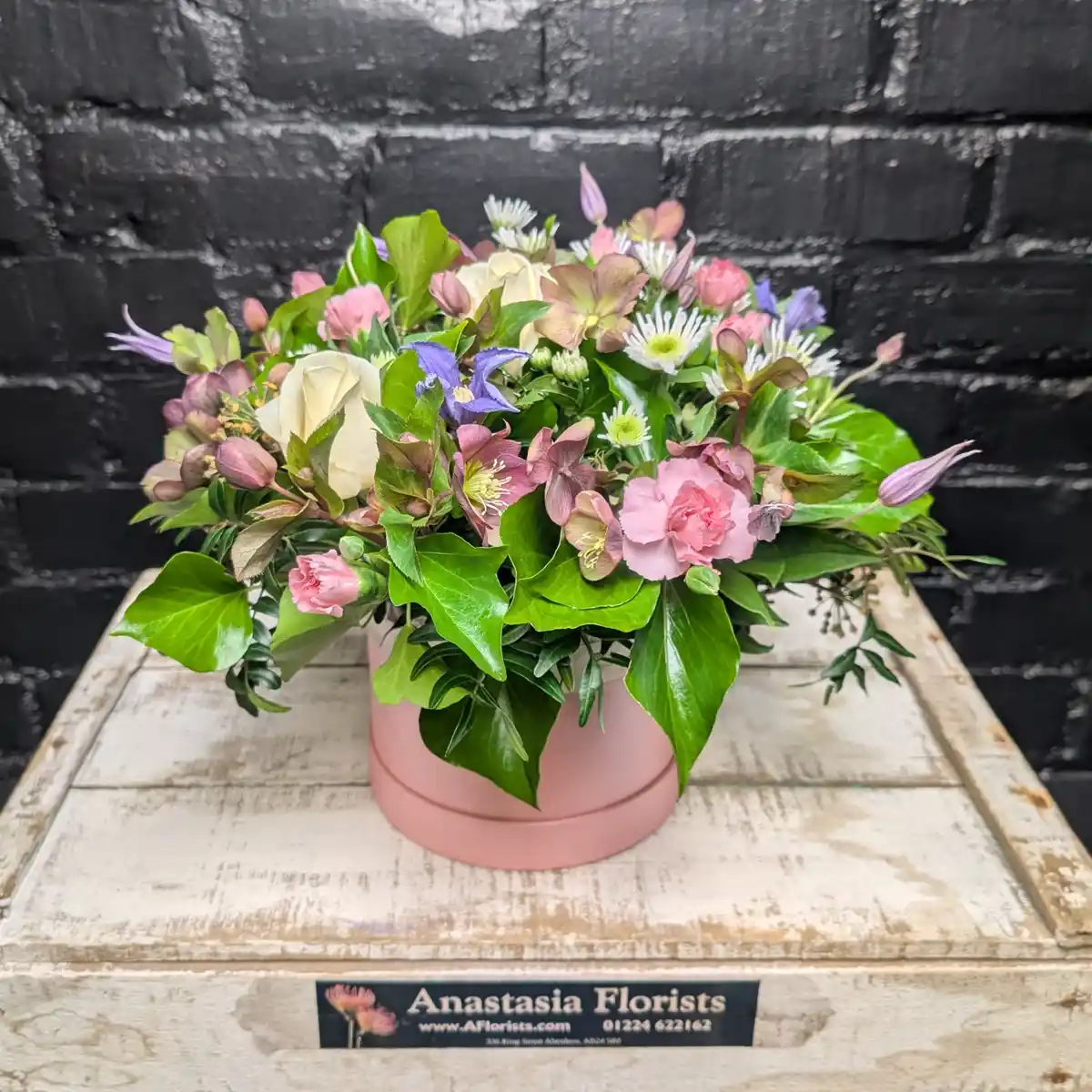 The Blush Garden Hat Box Far View - Floral arrangement in a pink hat box with cream roses, pink carnations, purple flowers, and green ivy leaves.