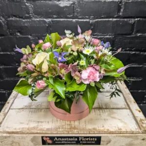 Floral arrangement in a pink hat box with cream roses, pink carnations, purple flowers, and green ivy leaves.