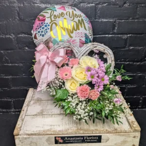 Heart-shaped wicker basket with pink roses, purple daisies, and a Love You Mum balloon for Mother's Day.