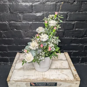 A tiered floral arrangement of white carnations, pink spray carnations, blue sea holly, and baby's breath in a white ceramic pot against a black brick wall.