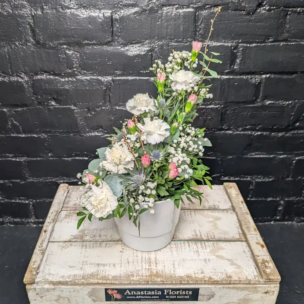 Mother’s Day Flower Pot A tiered floral arrangement of white carnations, pink spray carnations, blue sea holly, and baby's breath in a white ceramic pot against a black brick wall.