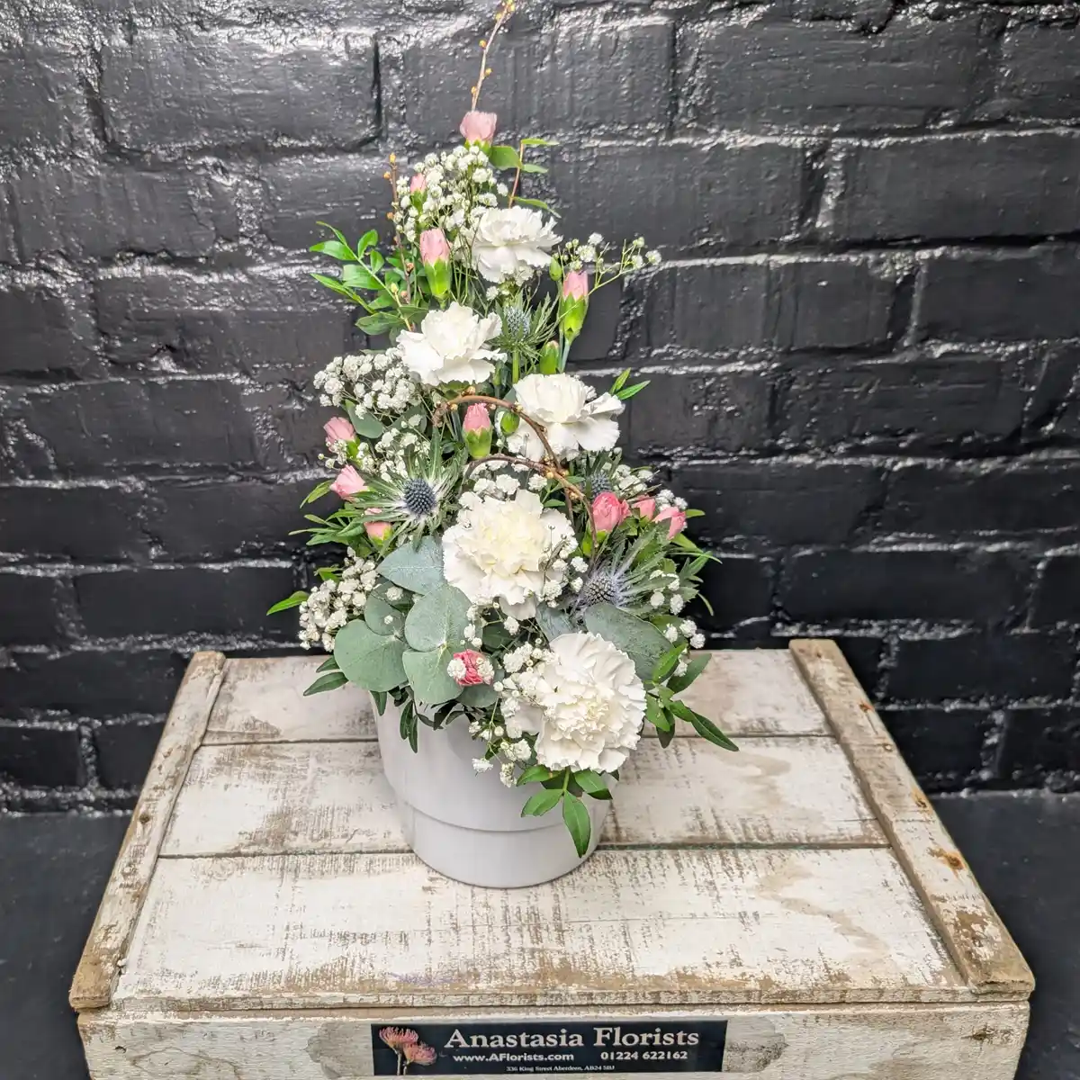 Mother’s Day Flower Pot Side View - A tiered floral arrangement of white carnations, pink spray carnations, blue sea holly, and baby's breath in a white ceramic pot against a black brick wall.