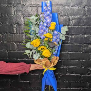 Person holding a tall blue delphinium and yellow rose graduation presentation bouquet with a miniature graduation cap against a black brick wall