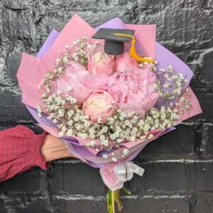 Hand holding a pink peony and baby's breath graduation posy tied with a white ribbon, topped with a mini graduation cap