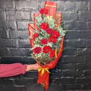 Florist in Aberdeen Person a tall red rose, carnation, and Germini graduation presentation bouquet with a miniature graduation cap against a black brick wall.