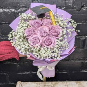 Hand holding a lilac rose and baby's breath graduation posy tied with a white ribbon, topped with a mini graduation cap.