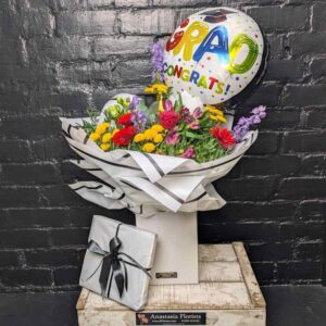 A vibrant graduation flower bouquet in a white presentation box, a "Grad Congrats" balloon, and a silver gift-wrapped box of chocolates against a black brick wall.