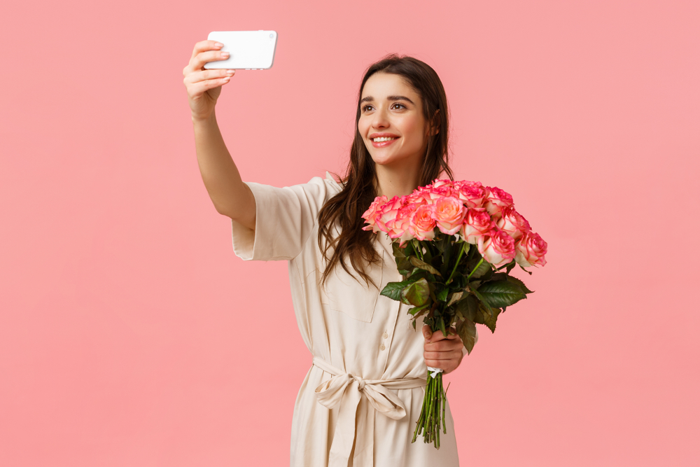 A woman takes a picture with flowers on a pink background